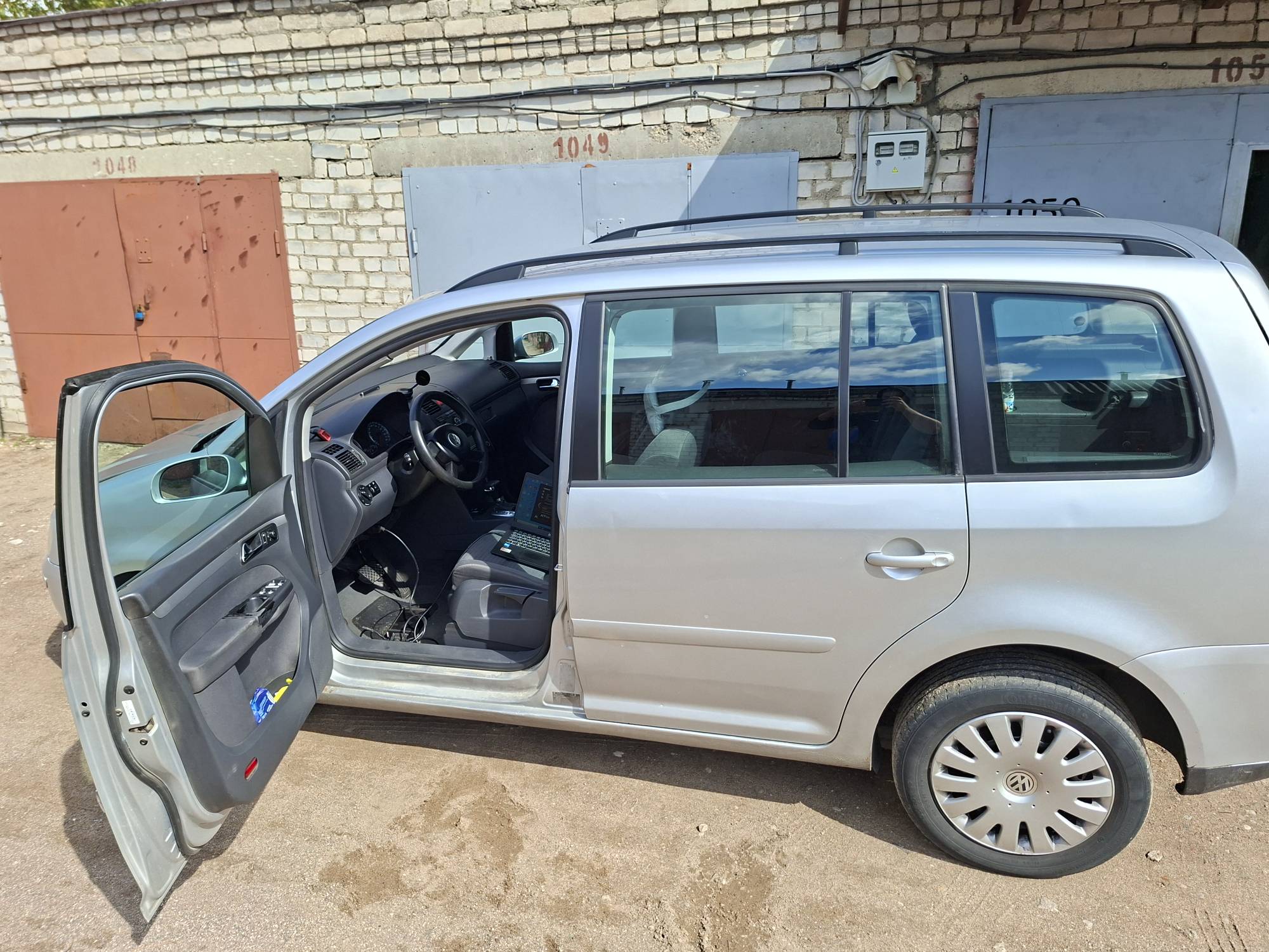 Серебристый универсал Volkswagen с открытой водительской дверью, внутри видно подключённый ноутбук для диагностики, автомобиль стоит у гаражей.