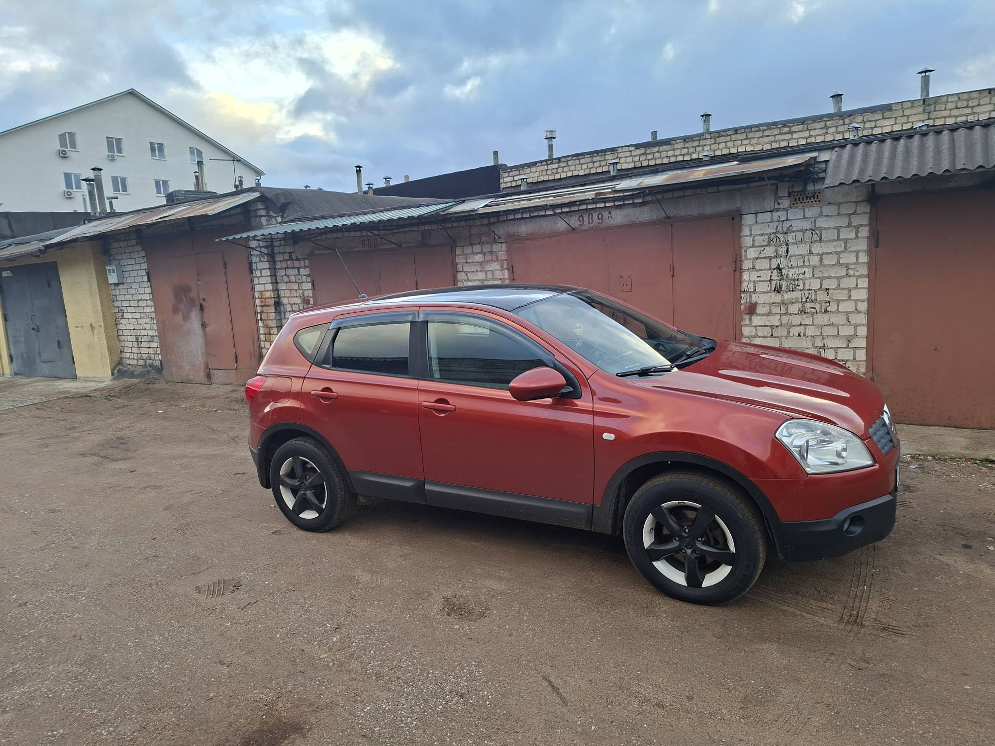 Красный Nissan Qashqai припаркован возле старых гаражей, вид сбоку, на заднем плане белый дом и облачное небо.